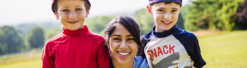 A woman smiles outdoors with two young boys, one in a red shirt and hat, the other in a blue hat and shirt that says “SNACK SHARK.” They are standing close together on a sunny day with greenery in the background.