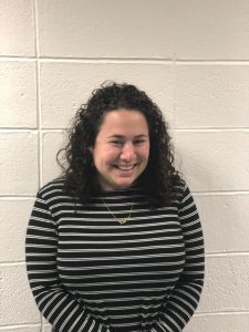 A person with curly dark hair, wearing a black and white striped long-sleeve shirt and a necklace, stands and smiles in front of a white textured wall with visible rectangular seams.