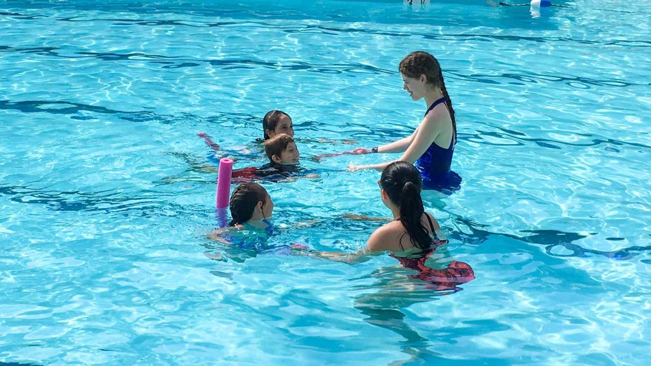 A swim instructor in a blue swimsuit teaches four children in a pool. The kids are gathered around her, some holding pool noodles. The water is clear and blue, and everyone appears to be engaged in the lesson.