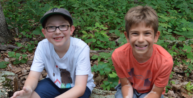 Two smiling boys sit outdoors on a forest floor with green leaves and fallen branches around them. One boy wears glasses, a cap, and a white shirt; the other wears an orange shirt. Both appear happy and playful.