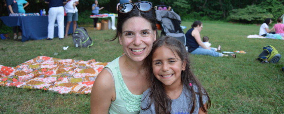 A woman and a young girl smile at the camera while sitting on grass at an outdoor gathering. People, blankets, and backpacks are visible in the background on a sunny day.