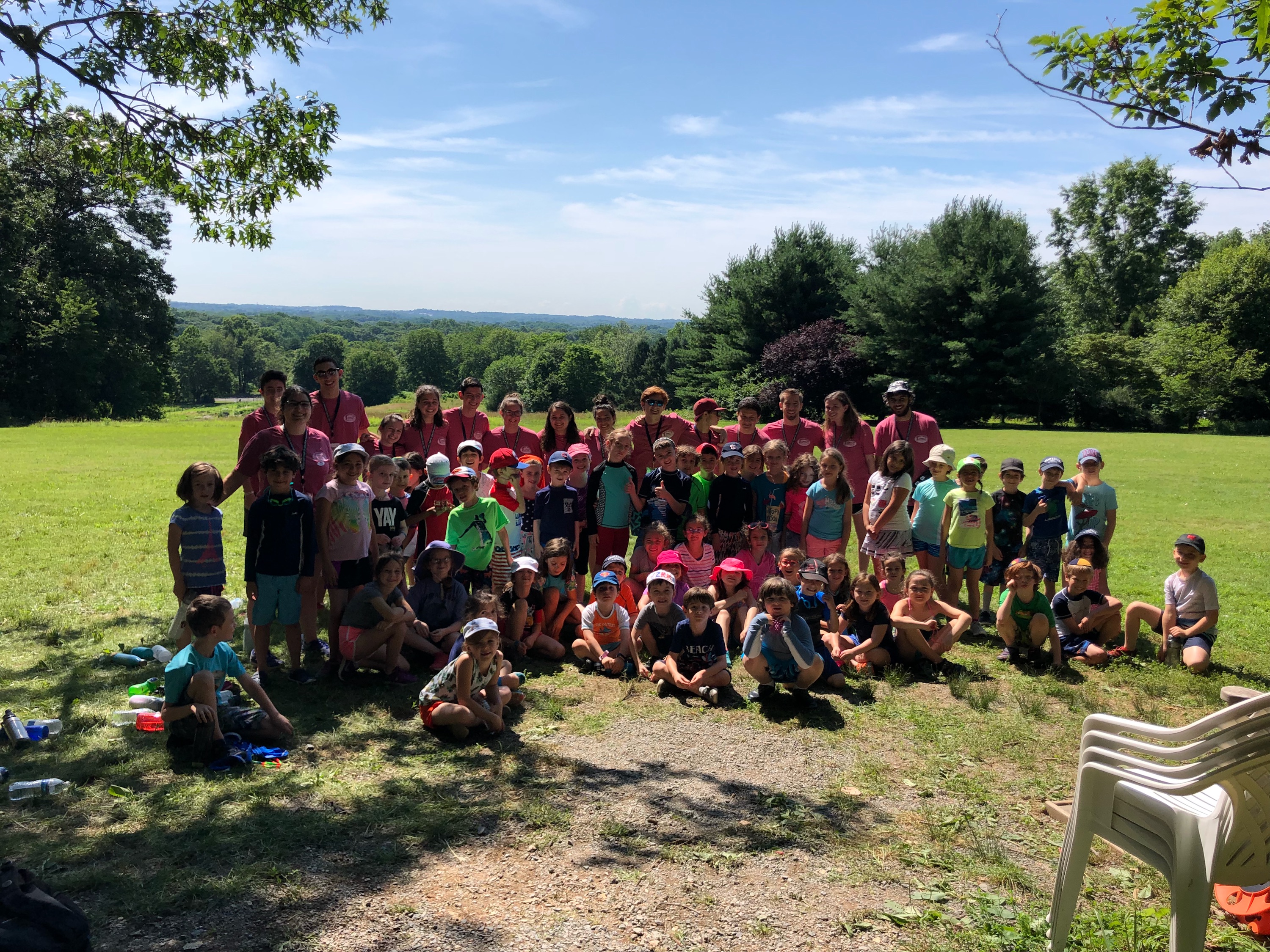 A large group of children and adults pose for a photo outdoors on a sunny day, surrounded by green grass and trees, with hills in the background. Several white plastic chairs are stacked on the right.