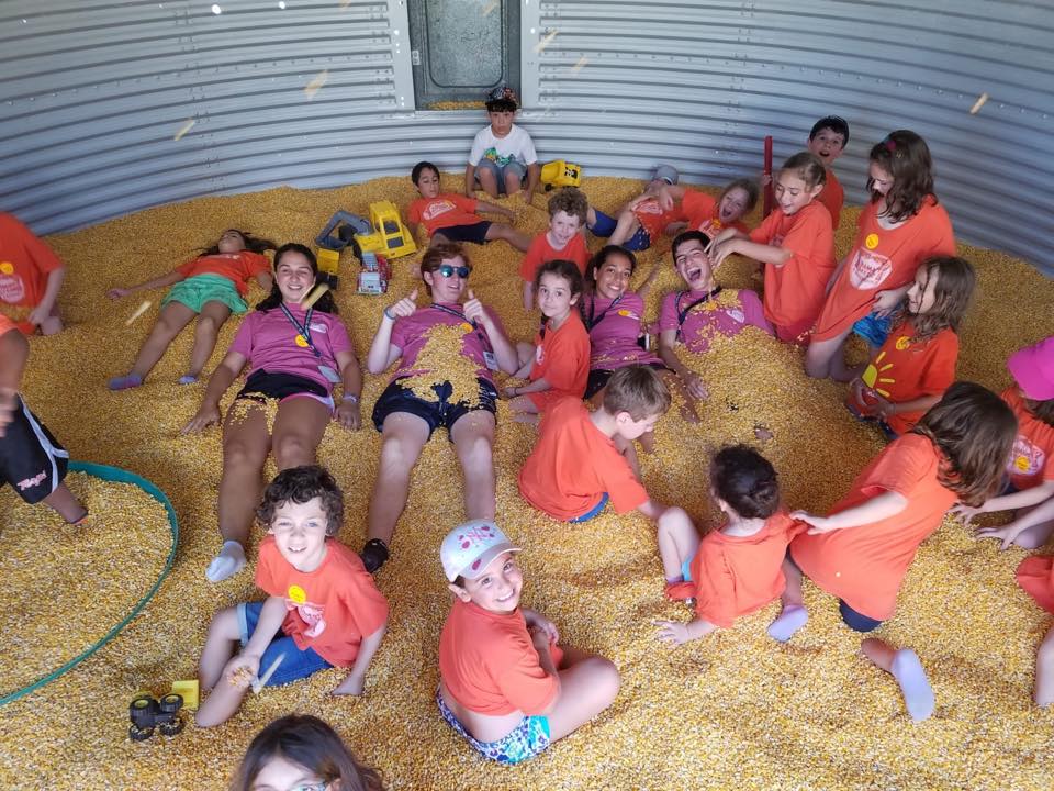 A group of children wearing orange shirts play and relax in a large container filled with corn kernels, smiling and laughing together. Some children have toy trucks and others are lying down, enjoying the playful environment.