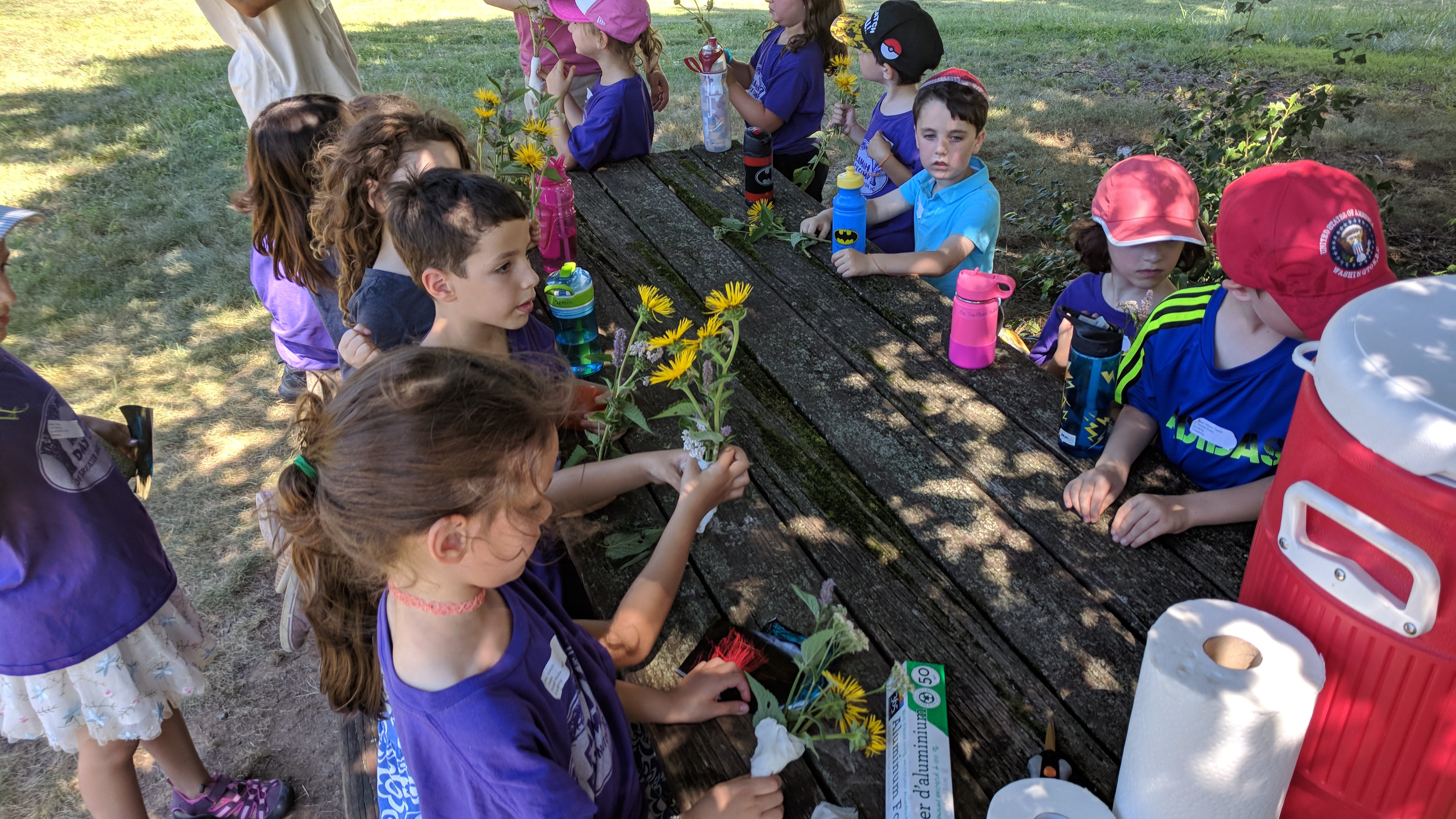 A group of children sit around a wooden picnic table outdoors, making small bouquets with yellow flowers. Water bottles and supplies are scattered on the table. The children are wearing colorful shirts and hats.