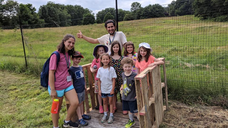 A group of children and two adults stand and smile together outdoors near a wooden bridge, with a grassy field and trees in the background. The sky is cloudy and everyone looks happy.