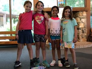 Four young girls stand side by side indoors, smiling at the camera. They wear casual summer outfits, and the background features a stone fireplace and large windows with trees visible outside.